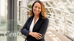 Aigerim Rachimow, freundlich lächelnd im Business-Outfit mit verschränkten Armen, steht in einem modernen, lichtdurchfluteten Bürogebäude.